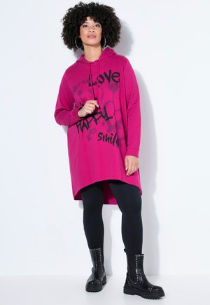 Frau mit lockigem Haar, die ein magentafarbenes Kapuzenkleid mit schwarzem Text, schwarze Leggings und schwarze, mit Nieten besetzte Stiefel trägt, vor einem weißen Hintergrund steht.