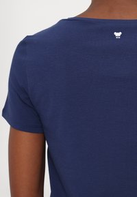 Camiseta de algodón azul marino con mangas cortas, con un escote redondeado y un pequeño logo blanco cerca de la parte trasera del escote. Textura suave.