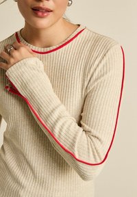 Beżowy, prążkowany sweter z długim rękawem z czerwonym wykończeniem przy kołnierzu i rękawach. Posiada dopasowany krój oraz miękką fakturę.