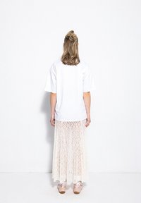 Biała, oversize t-shirt zestawiona z długą, przezroczystą spódnicą koronkową w kolorze kremowym. Modelka nosi jasnoróżowe buty z tasiemkami na kostkach.