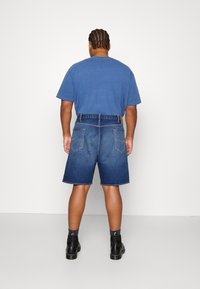 Pantalones cortos de mezclilla en azul oscuro con corte recto, que cuentan con dos bolsillos traseros y una etiqueta de cuero en la cintura. Combinados con una camiseta azul.