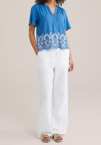 Blå blus med vit broderad fåll, korta ärmar och v-ringning; tillsammans med vita linnebyxor och beigea slip-on sandaler.