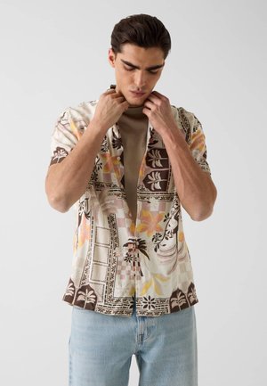Homme ajustant le col d'une chemise à manches courtes ouverte avec des motifs floraux et géométriques sur un t-shirt marron uni, portant un jean bleu clair.