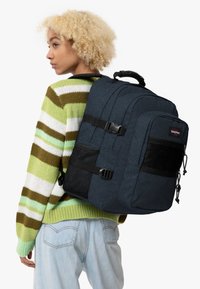Ung person med lockigt blont hår som bär en randig grön tröja och ljusblå jeans, bär en stor marinblå ryggsäck på axeln.