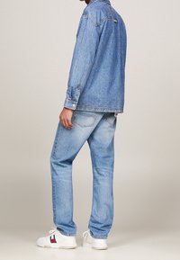 Man wearing light blue denim jacket and jeans with white sneakers, standing against plain beige background.