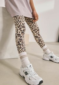 Beige és barna leopárdmintás leggings, testhez simuló dizájnnal; fehér New Balance edzőcipővel és csíkos zoknival párosítva.