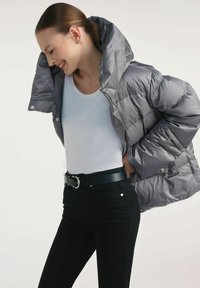 Szara kurtka puchowa z wysokim kołnierzem, pikowanym designem i błyszczącym wykończeniem, noszona na białej koszuli i czarnym pasku, w połączeniu z czarnymi dżinsami.