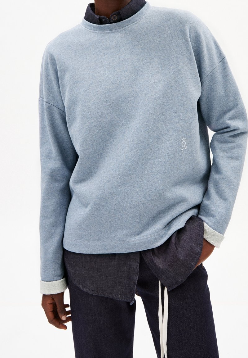 Personne portant une chemise bleu clair à manches longues par-dessus une chemise sombre boutonnée avec un pantalon sombre, une main dans la poche sur un fond uni.
