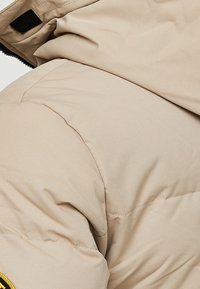 Sección de chaqueta acolchada beige que muestra una textura acolchada, tela elástica y cierre de Velcro negro. Presenta costuras a lo largo de las uniones.