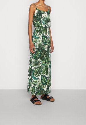 Kobieta w długiej sukience na ramiączkach z zielonym tropikalnym wzorem liści oraz czarnych sandałach z podwójnymi paskami, stojąca przy białej ścianie.