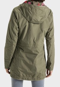 Personne portant une veste à capuche longue à manches longues de couleur vert olive avec une doublure rose à motifs, vue de dos.