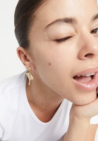 Pendientes de tono dorado con un diseño escultórico que recuerda a una hoja o una vela, con una superficie lisa y reflectante y un adorno colgante.