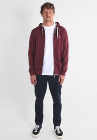 Sweat à capuche zippé bordeaux avec cordons, porté sur un t-shirt blanc, associé à un pantalon bleu marine et des baskets noires. Design simple, coupe décontractée.