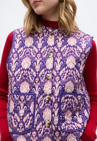 Γυναίκα που φοράει γιλέκο χωρίς μανίκια με κουμπιά και ύφασμα σε μοτίβο μωβ και ροζ πάνω από κόκκινο μπλουζάκι με μακριά μανίκια, με τα χέρια στις τσέπες του γιλέκου.