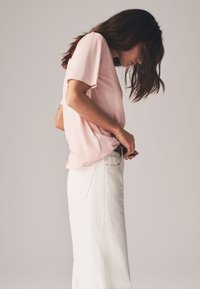 Ljusrosa bomulls-T-shirt med korta ärmar, kombinerad med högmidjade vita denimjeans med fickor, stylad i profil.