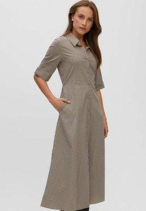 Woman with long hair wears a beige checkered button-up dress with half sleeves, posing with one hand in pocket against a plain background.