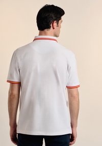 Camisa polo blanca con cuello y acentos en las mangas de rayas rojas y amarillas, con una textura suave y mangas cortas. Vista trasera mostrada.