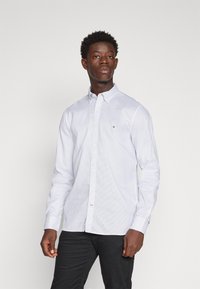Camicia bianca a maniche lunghe con un piccolo motivo blu, colletto button-down e un dettaglio logo sul petto, abbinata a pantaloni neri.