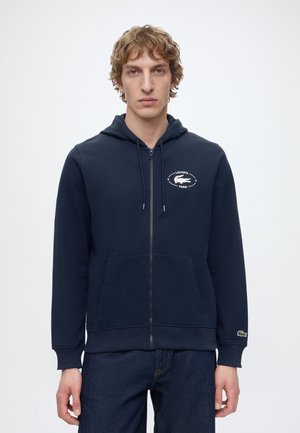 Man draagt een marineblauwe hoodie met rits en het Lacoste-logo op de borst en mouw, staand tegen een effen lichte achtergrond.