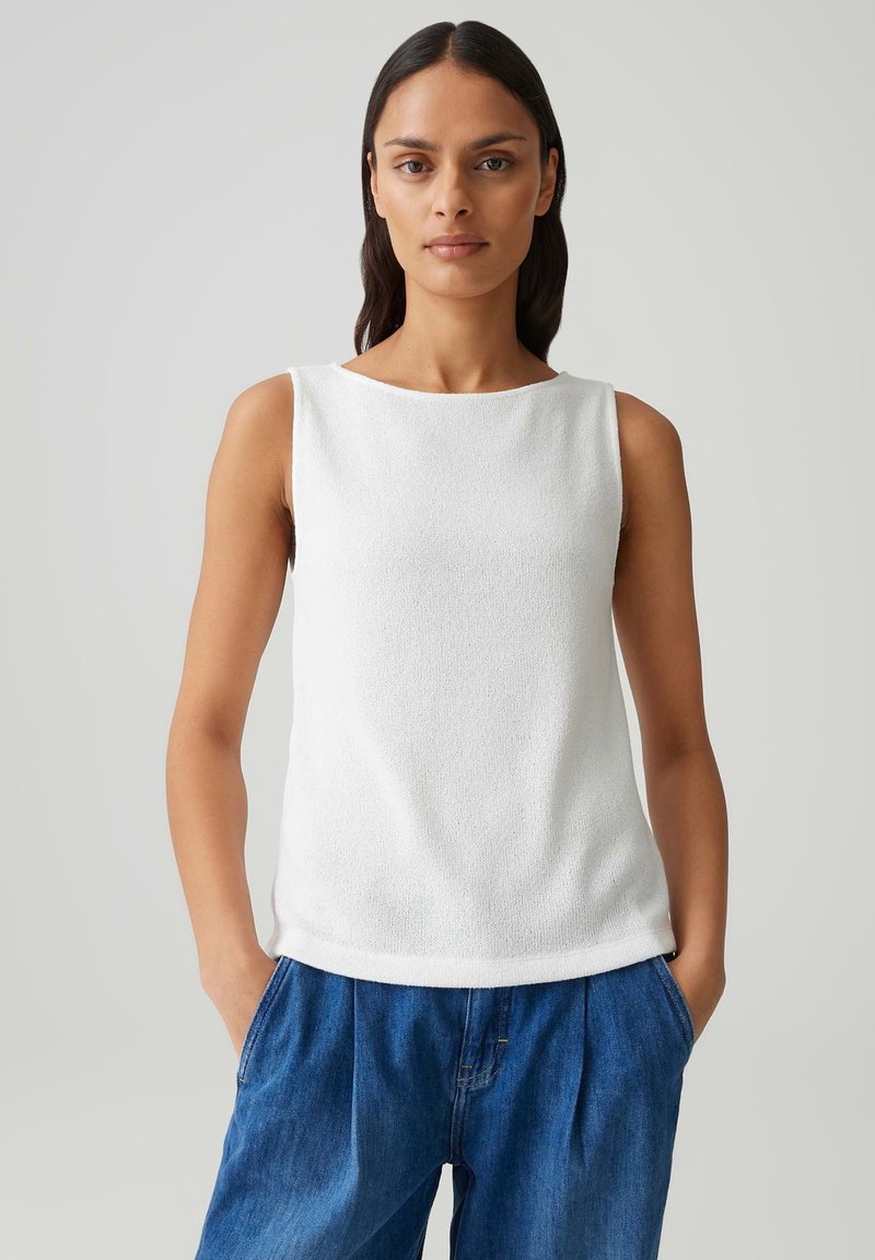 Donna con lunghi capelli scuri che indossa un top bianco senza maniche e pantaloni di denim blu larghi, con le mani in tasca, in piedi davanti a uno sfondo grigio.