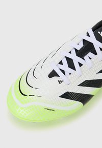 Chaussure de football blanche avec une tige texturée, accents noirs et une semelle vert fluo. Présente des motifs rayés et des lacets blancs.
