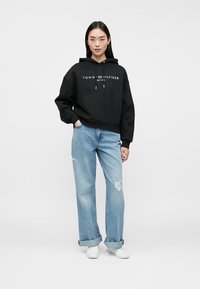 Juodas trumpas gobtuvėlis su baltu "TOMMY HILFIGER" logotipu, derinamas su šviesiai mėlynomis nusidėvėjusiomis džinsomis ir baltais sportbačiais. Paprasta, laisvalaikio siluetas.