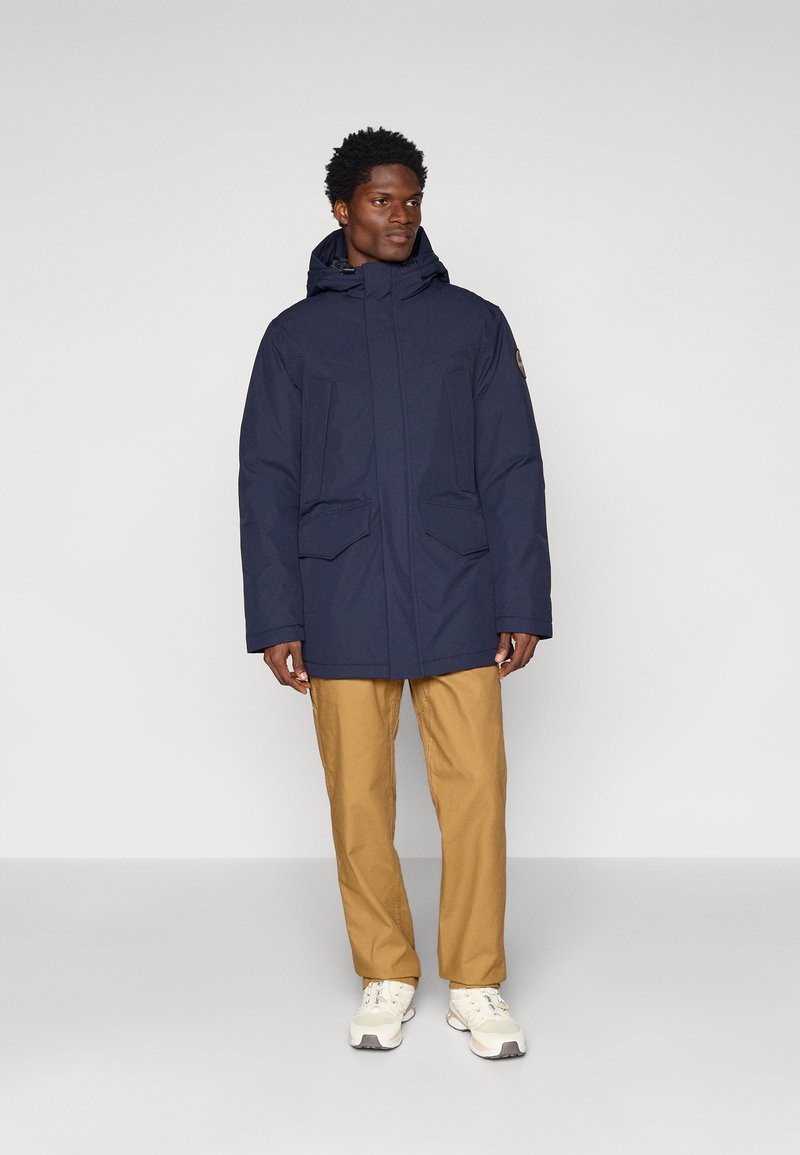Veste imperméable navy avec deux poches avant, un col montant et une capuche attachée, associée à un pantalon beige et des baskets blanches.
