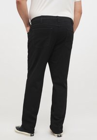 Pantalones de mezclilla negros con diseño de corte recto, cinco bolsillos y costuras en contraste. Combinados con zapatos oscuros y una camisa blanca sencilla.