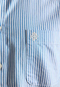 Niebiesko-biała koszula w pionowe paski z kieszenią na piersi, z białymi haftowanymi inicjałami nad kieszenią.