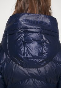 Mörkblå puffjacka med glansig yta, som har huva och kviltad design. "Calvin Klein"-logotyp präglad på baksidan.