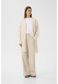 Cardigan bej tricotat cu mâneci lungi și cu față deschisă, purtat peste o cămașă albă și pantaloni bej cu croială largă, asortat cu sandale negre.