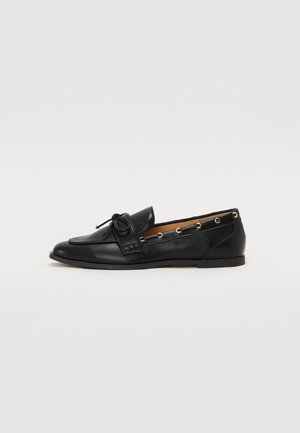 Mocassin plat en cuir noir avec un détail nœud sur le dessus et des œillets dorés sur le côté, sur un fond clair uni.
