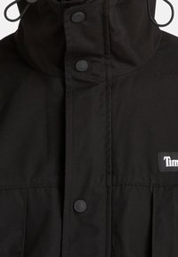 Svart jacka med hög krage, tryckknappar och en logotyp patch. Har en slät yta och en strukturerad design för funktionalitet.