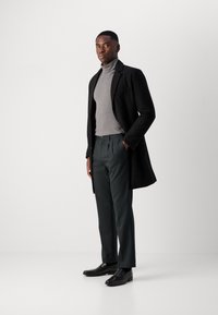 Manteau noir, col roulé gris, pantalon vert et chaussures noires. Tissu lisse avec un design épuré, coupe ajustée et détails subtils.