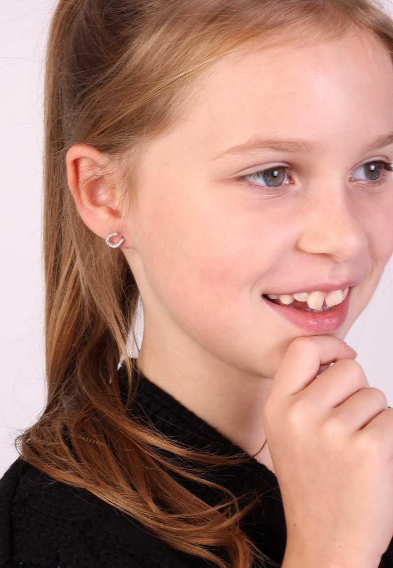 Silberne Ohrstecker in Herzform mit einer glatten Metallic-Oberfläche. Ein Kind mit langen, hellbraunen Haaren und einem schwarzen Kleidungsstück wird gezeigt.