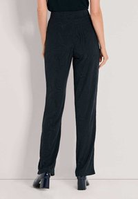 Vrouw in hoogwagende zwarte vlakke stoffen broek met een subtiele golvende patroon, gecombineerd met zwarte blokhakken, staand tegen een effen achtergrond.