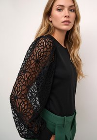 Black top with lace puff sleeves featuring a floral pattern. Paired with green high-waisted pants, showcasing a textured fabric.