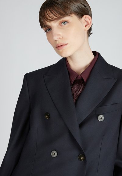 Blazer doppiopetto blu navy in tessuto strutturato con ampi revers, bottoni metallici e taschino sul petto, indossato sopra un top bordeaux.