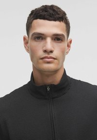 Juodas zip-up džemperis su aukštu apykakle, dekoruotas tekstūruotu mažu baltų taškų raštu. Standartinė forma, lengvas audinys.