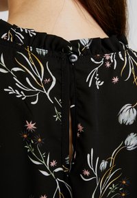 Blouse noire avec motif floral multicolore, col volanté et une seule boutonnière noire au niveau de la nuque.