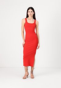 Red sleeveless ribbed midi dress with a square neckline and side slit. Model wears beige strappy heels showcasing a tattoo on the ankle.