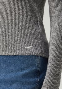 Grå stripet genser laget av teksturert stoff, med en liten hvit logo nederst, kombinert med blå denimjeans.