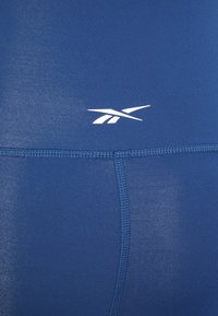 Tecido atlético azul com uma textura lisa e um logótipo da Reebok em branco. Apresenta costura em contraste e um design ajustado.
