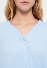 Ανοιχτό γαλάζιο ένδυμα με V-λαιμόκοψη και υφή υφάσματος. Διαθέτει κουμπιά κατά μήκος της μπροστινής πλευράς, προβάλλοντας μια λεία και απαλή εμφάνιση.