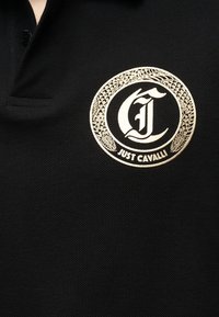 Μαύρο πόλο πουκάμισο με χρυσλό κυκλικό λογότυπο Just Cavalli που περιλαμβάνει ένα στυλιζαρισμένο "JC" και διακοσμητικά στοιχεία φιδιού στο στήθος.