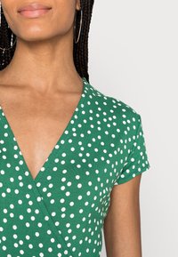 Zielona bluzka w kropki z krótkimi rękawami i głębokim dekoltem w kształcie V. Wykonana z gładkiego, lekkiego materiału o miękkiej fakturze.