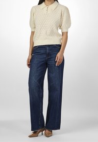 Maglione in cotone lavorato a maglia con pattern a diamante e dettaglio a bottoni, abbinato a jeans a gamba larga blu scuro e décolleté a punta color marrone chiaro.
