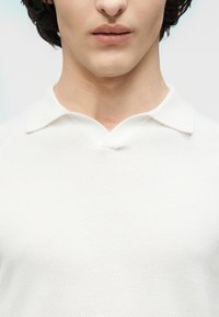 Jeune personne portant un polo en maille blanche texturée avec un col en V, visible de la mâchoire à la poitrine sur un fond uni.