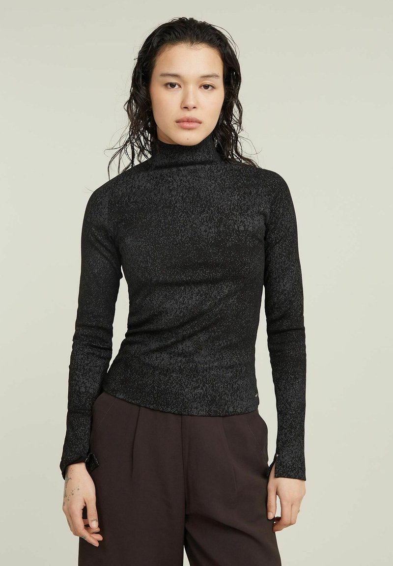 Kobieta z mokrymi włosami, ubrana w dopasowany czarny golf z długim rękawem i luźne ciemno-brązowe spodnie, stojąca na tle jasnym jednolitym.