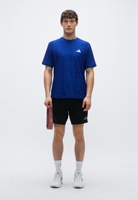 T-shirt de sport bleu à motif texturé, short noir et baskets blanches. Tenant un élastique de résistance rouge dans la main gauche.